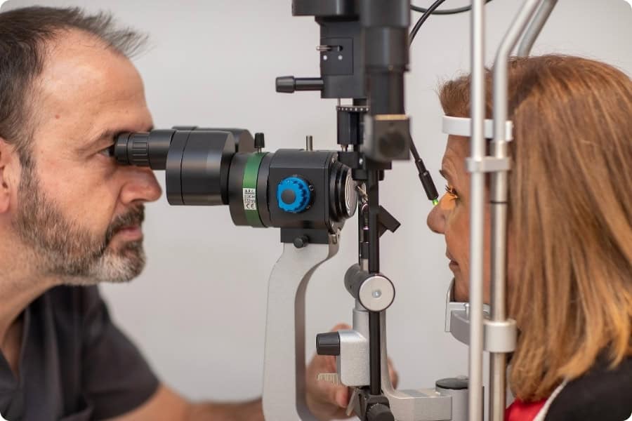 Un hombre con barba examina con precisión el ojo de una mujer con una lámpara de hendidura en el consultorio de un optometrista, con la vista puesta en posibles problemas quirúrgicos en la retina. El equipo se interpone entre ellos mientras el pelo castaño de ella enmarca la escena. Un hombre con barba examina con precisión el ojo de una mujer con una lámpara de hendidura en el consultorio de un optometrista, con la vista puesta en posibles problemas quirúrgicos en la retina. El equipo se interpone entre ellos mientras el pelo castaño de ella enmarca la escena.