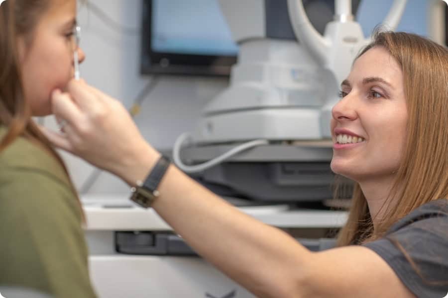 Una mujer con el pelo largo sonríe mientras examina la vista de un niño que lleva una camiseta verde. El niño está sentado frente a un equipo en el consultorio de un optometrista. El foco está puesto en la interacción entre la mujer y el niño. Una mujer con el pelo largo sonríe mientras examina la vista de un niño que lleva una camiseta verde. El niño está sentado frente a un equipo en el consultorio de un optometrista. El foco está puesto en la interacción entre la mujer y el niño.