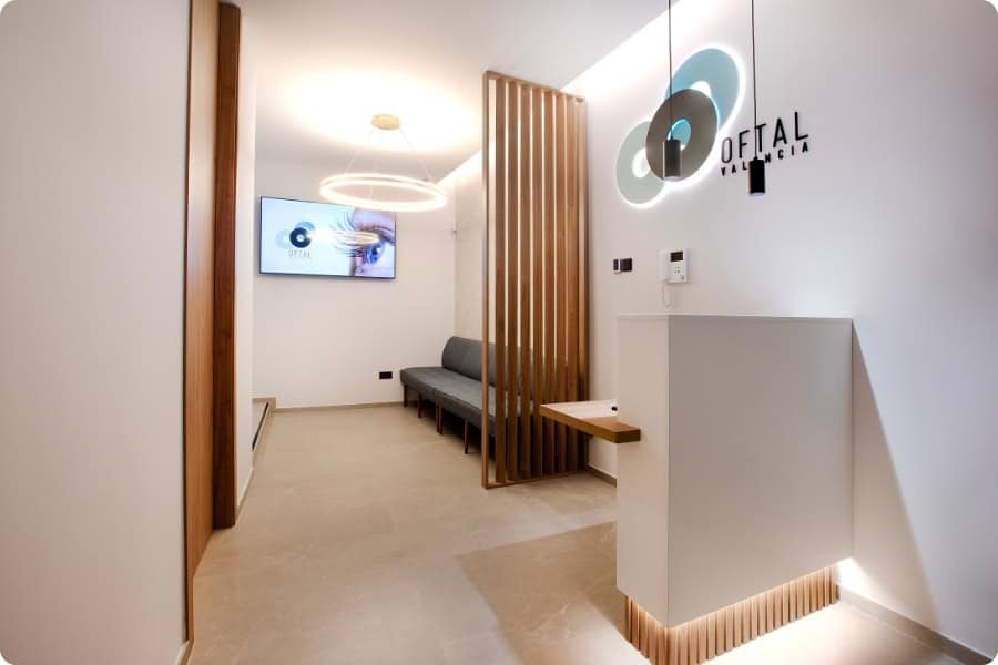 Sala de espera luminosa y moderna con detalles en madera, un banco acolchado a lo largo de la pared y un mostrador de recepción blanco. Una lámpara circular cuelga del techo. Una pantalla de televisión en la pared muestra "OFTAL", junto con información sobre PRP (Plasma Rico en Plaquetas). Sala de espera luminosa y moderna con detalles en madera, un banco acolchado a lo largo de la pared y un mostrador de recepción blanco. Una lámpara circular cuelga del techo. Una pantalla de televisión en la pared muestra "OFTAL", junto con información sobre PRP (Plasma Rico en Plaquetas).